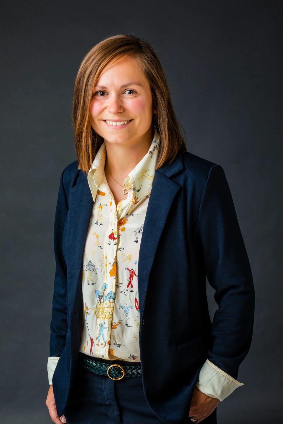 Portrætfoto af smilende kvinde i mørkeblå blazer og farvet skjorte, armene på hoften, mod grå baggrund.