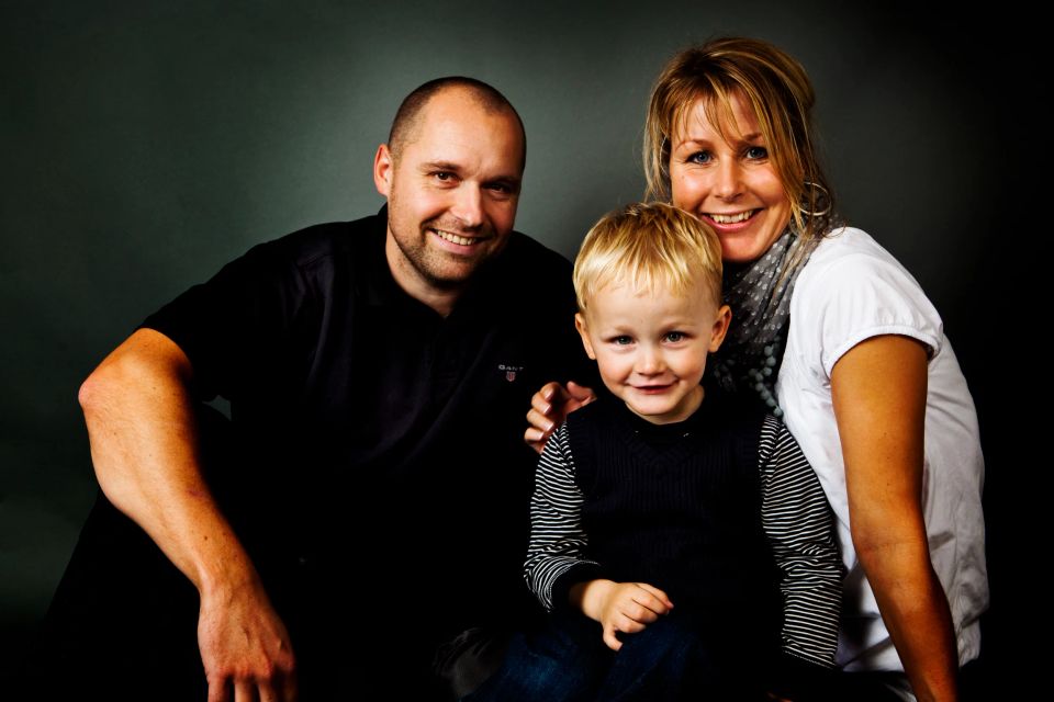 Familiefoto med smilende forældre og deres lille søn, naturligt lys og mørk baggrund.