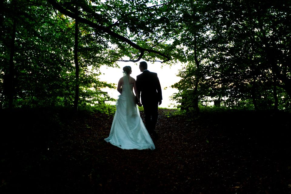 Bryllupsfoto af brudepar i silhuet, der går hånd i hånd ad skovsti med lys for enden. Stemningsfuld fotografering.