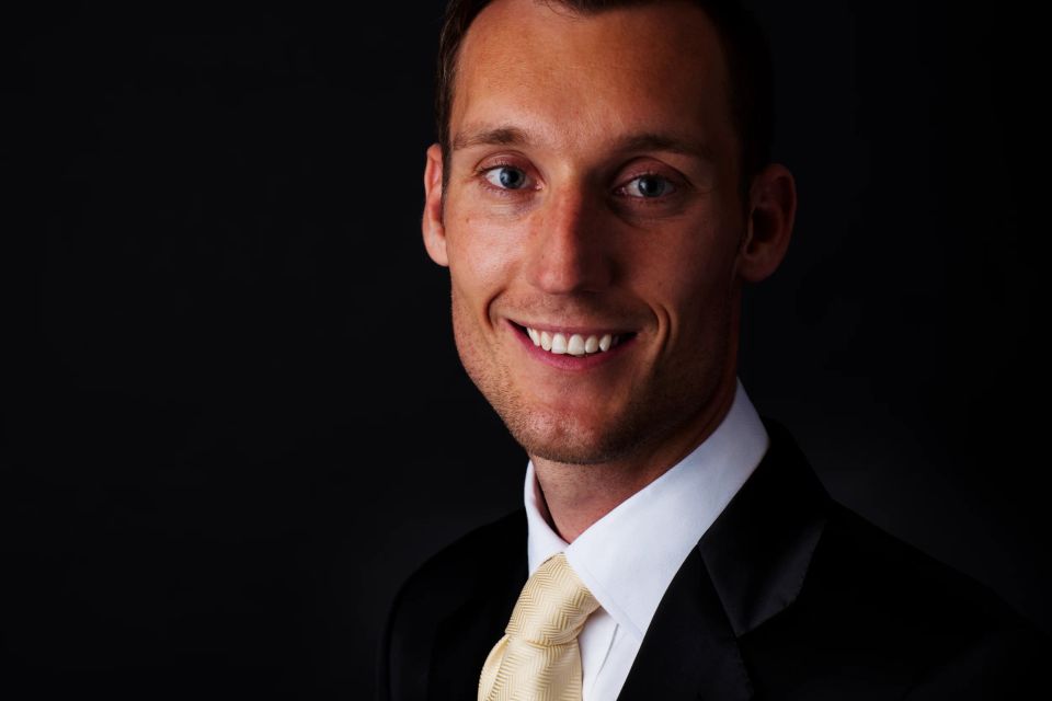 Professionelt portrætfoto af smilende mand i sort jakkesæt med guldbind, belyst fra siden mod mørk baggrund.