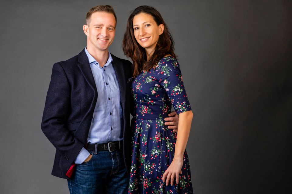 Glad portrætfotografering af et par, der smiler varmt, kvinden i blomstret kjole og manden i jakke på neutral baggrund.