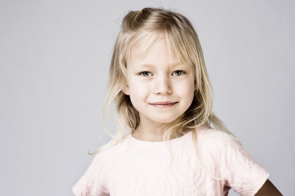 Børnefotografi af en ung pige med lyst hår og grønne øjne, der smilende kigger direkte ind i kameraet.