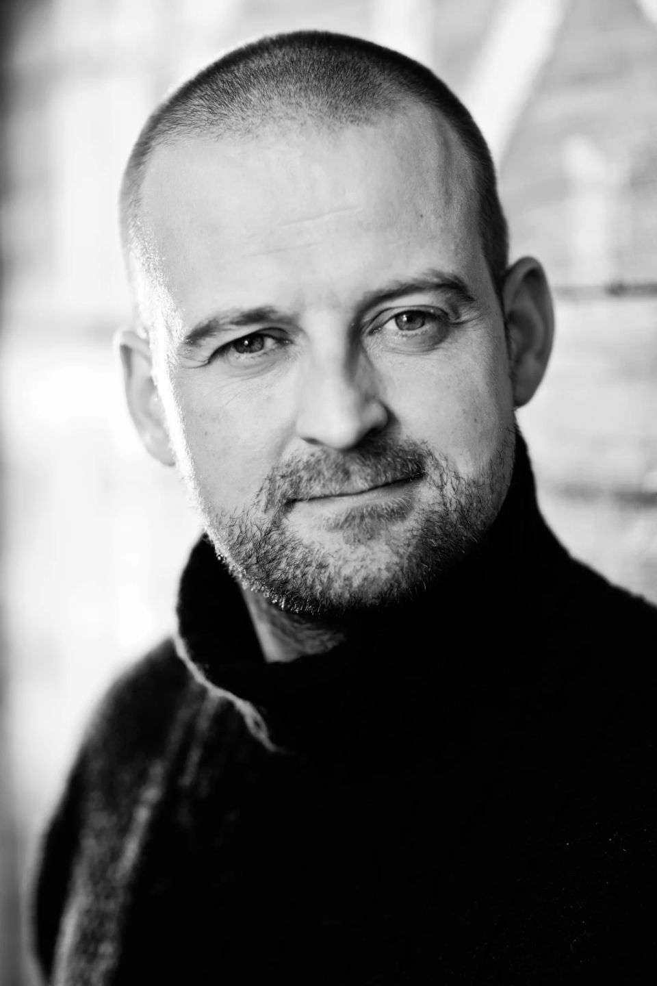 Professionelt sort-hvid portrætfoto af en smilende mand med skæg og kort hår i en sort rullekrave.