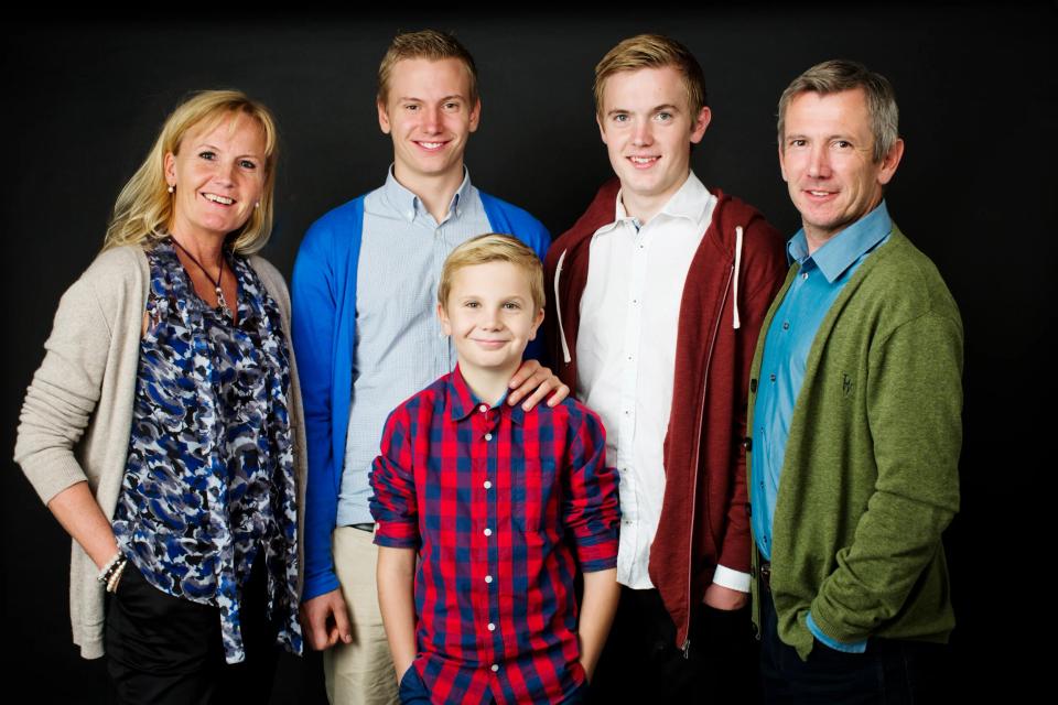Familie fotografering. Studieportræt af to forældre, to teenagesønner og en yngre søn mod en mørk baggrund.