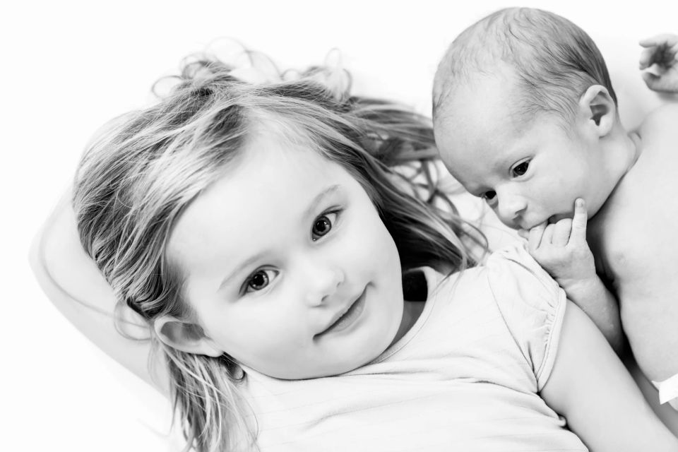Sort/hvid babyfotografering med storesøster, der smiler til kamera, mens lillebror putter sig kært ind til hende.