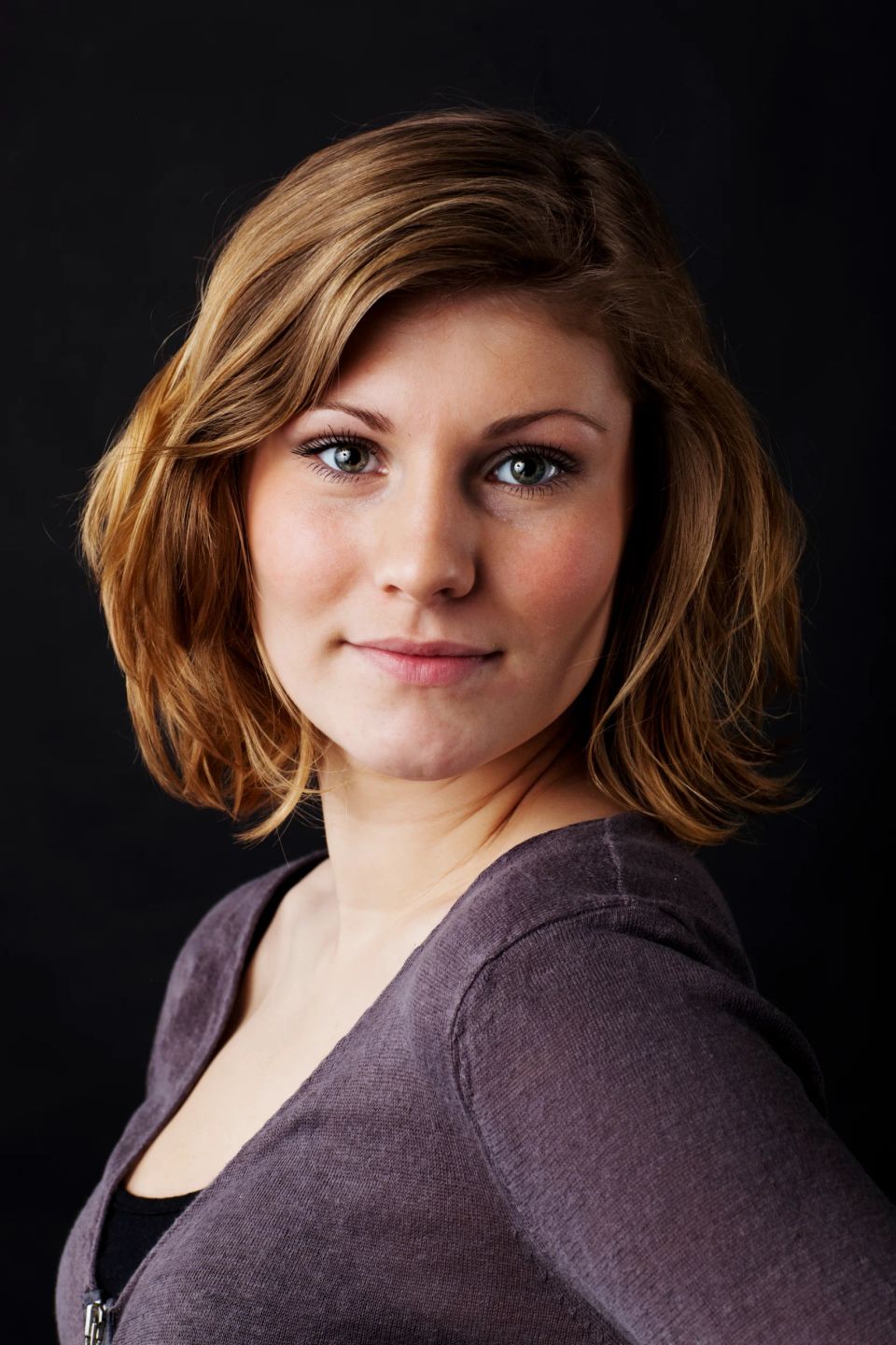 Nært portrætfoto af ung kvinde med brunt hår og gønne øjne, der smiler venligt, mod sort baggrund.