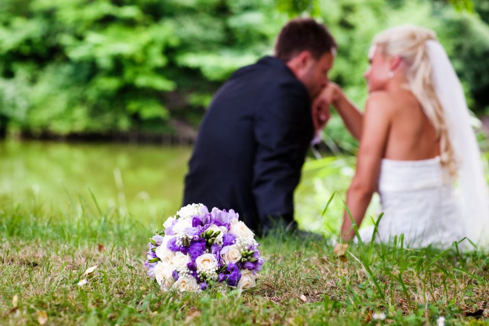 Bryllupsbillede af brudepar, der sidder i græsset ved en sø, med en smuk buket blomster i forgrunden.