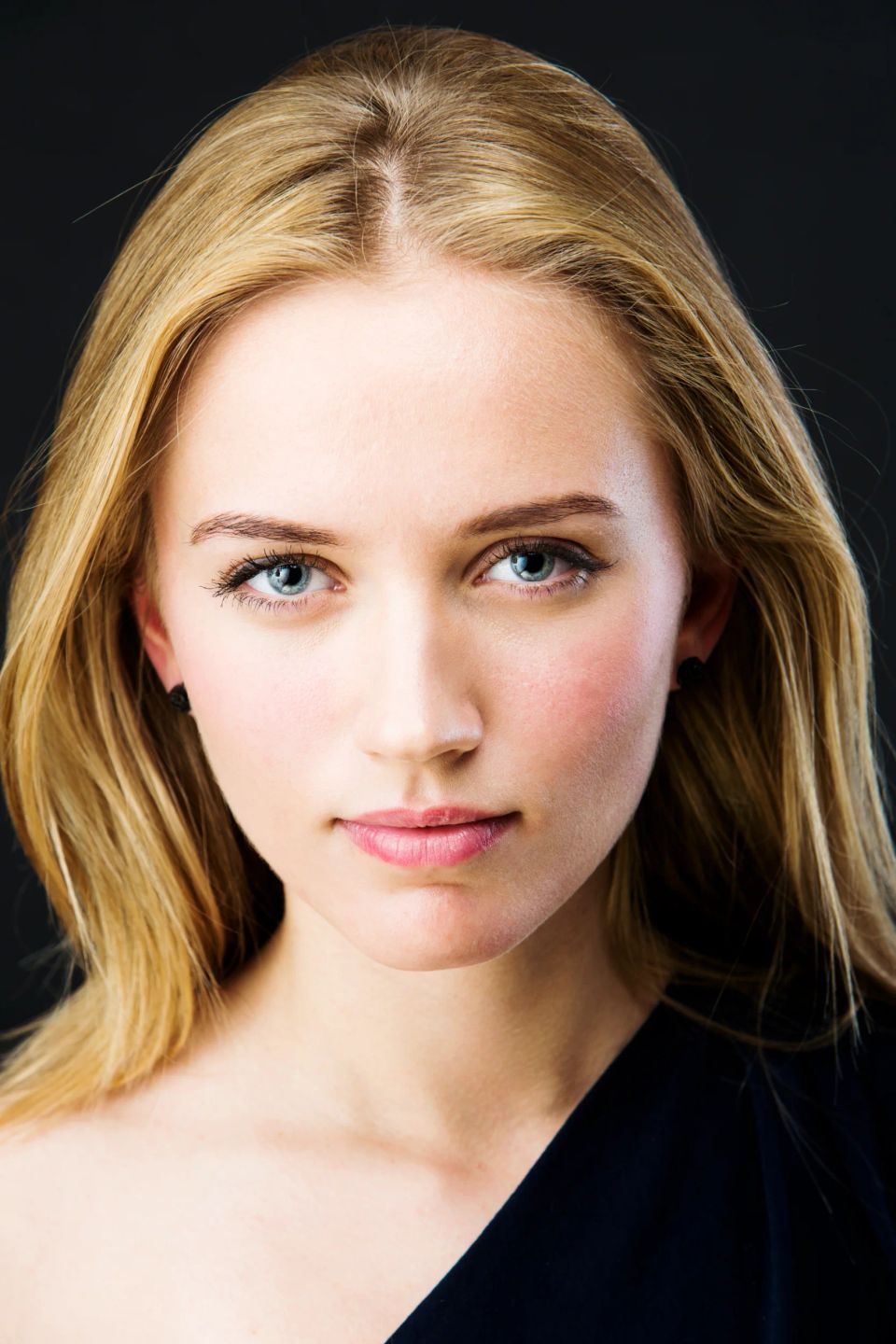 Nært portrætfoto af ung kvinde med blåt stirrende blikfangerblik, lyst hår og naturligt smil mod mørk baggrund.