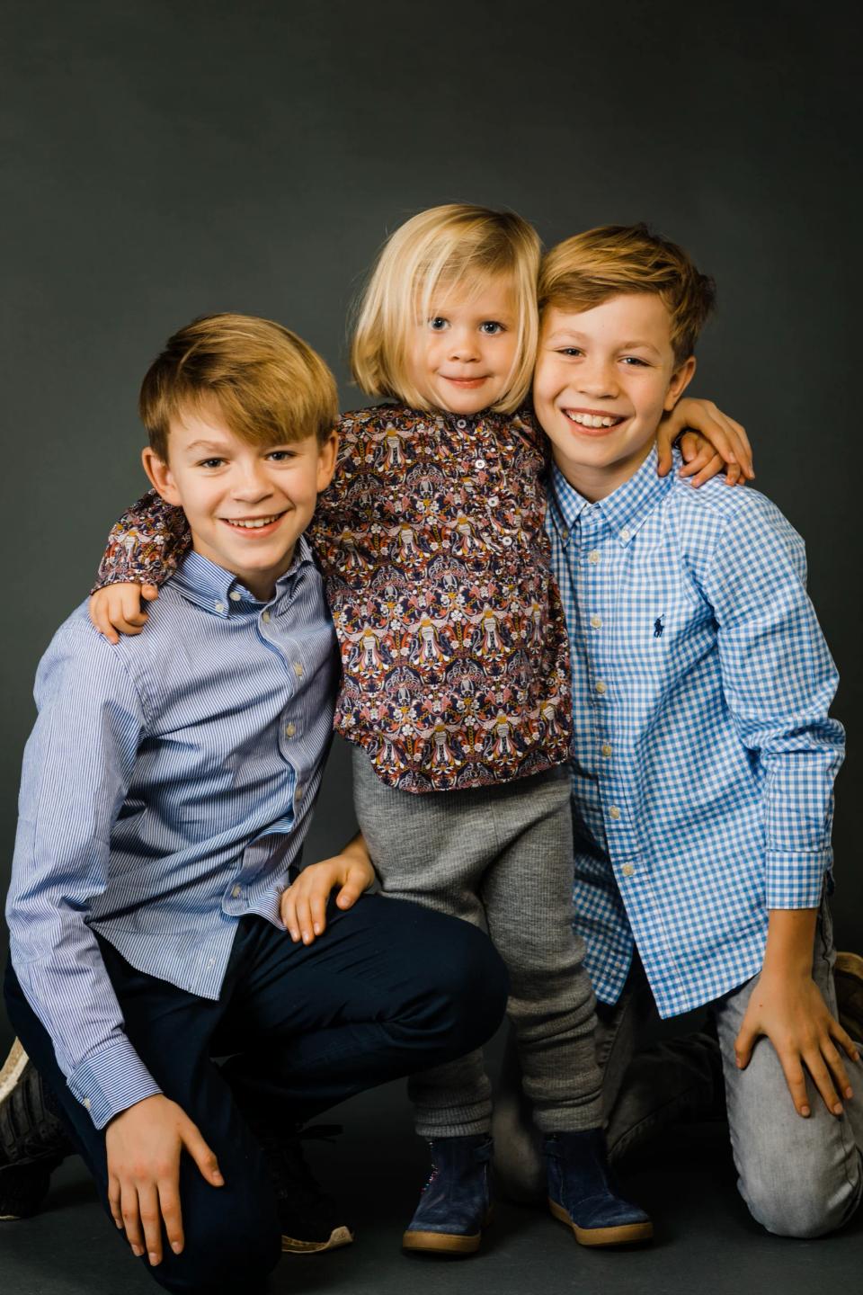 Familiefoto med tre smilende børn, to drenge og en lille pige, i kærlig omfavnelse mod grå baggrund. Et livligt familiepor...