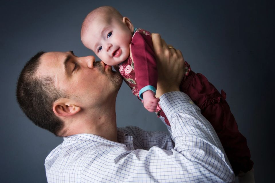 Babyfotografering: Nærbillede af far, der kysser sin baby, som smiler, med blød belysning mod mørk baggrund. Et autentisk ...