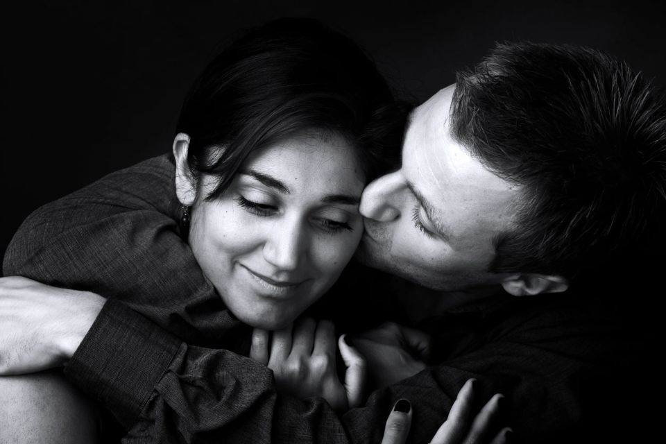 Sort/hvid portrætfoto af mand der kysser kvinde blidt på kinden. Begge er omsluttet af en intim, varm atmosfære.
