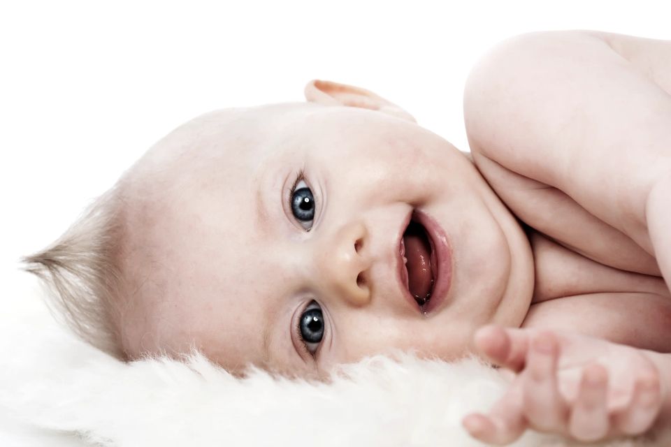 Glædeligt babyfotografi af smilende baby liggende på ryggen med åben mund og strålende blå øjne på hvidt tæppe.