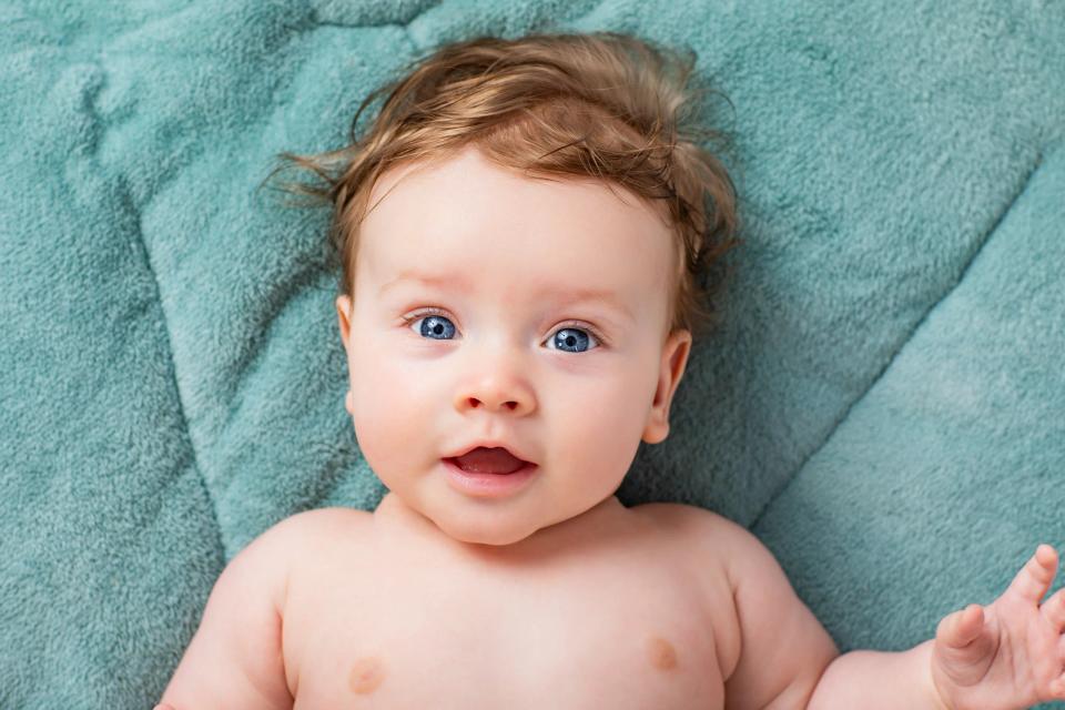 Glad baby med strittende brunt hår og klare blå øjne, som kigger direkte ind i kameraet fra et blåt tæppe.