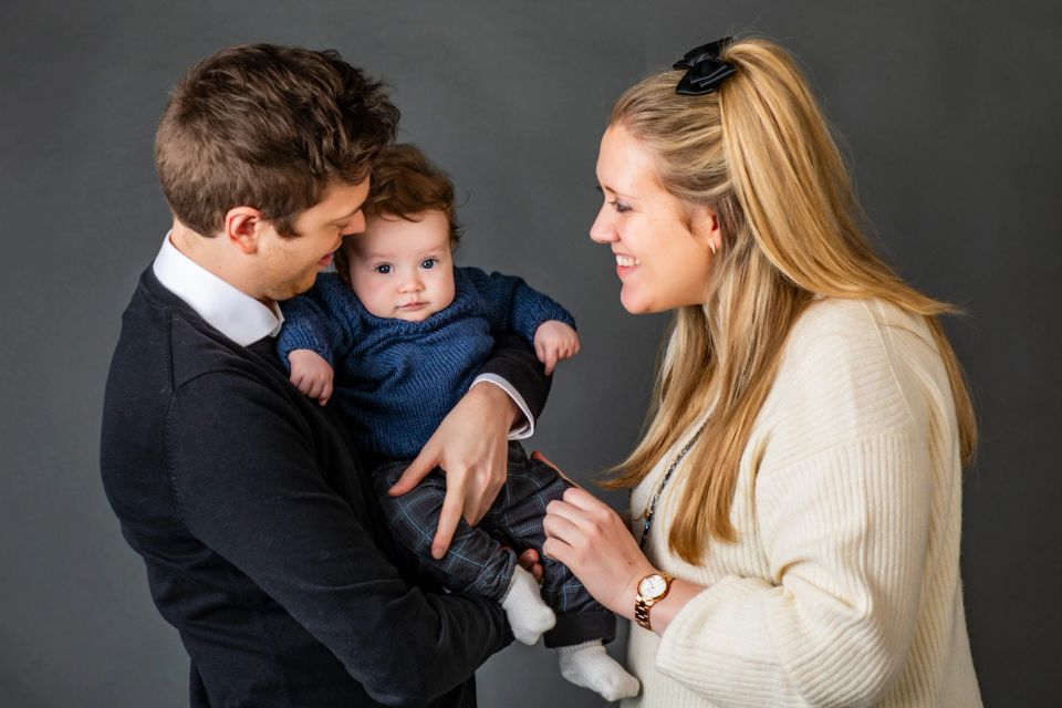 Familiefoto af smilende mor og far der holder deres baby i blødt studie-lys. Baby kigger direkte ind i kameraet.
