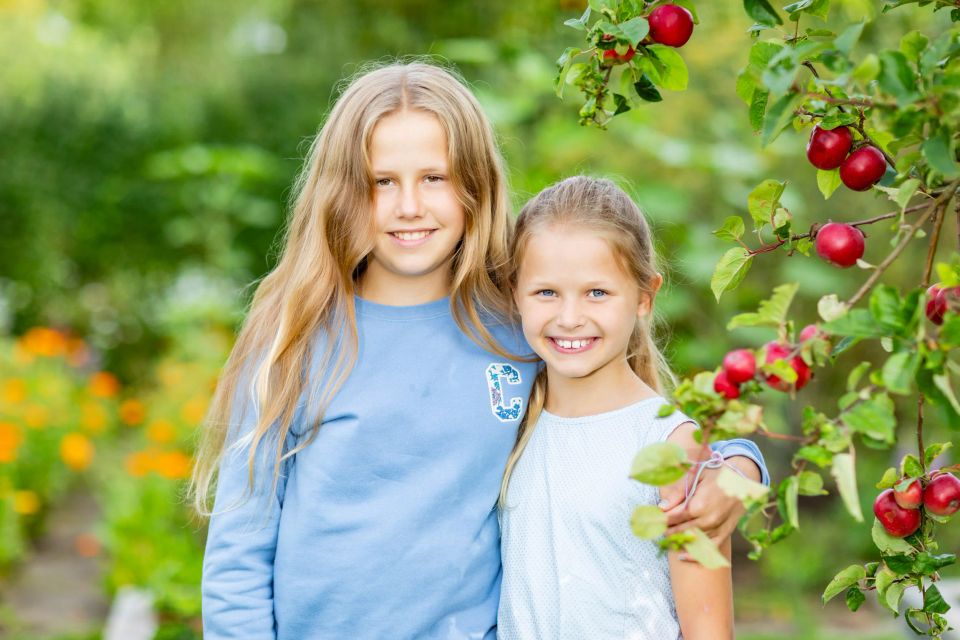 Børn: To smilende søstre i blå nuancer omfavner hinanden under et æbletræ i haven med grøn baggrund og røde æbler i forgru...