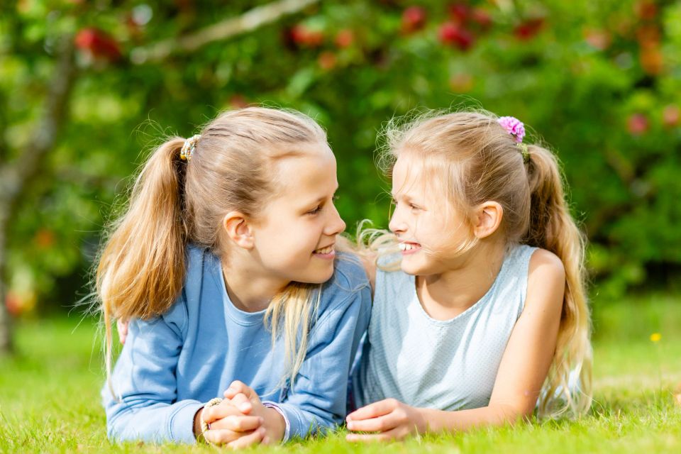 Legende børn: To piger med lyst hår smiler til hinanden, liggende i Virums grønne græs med sollys.