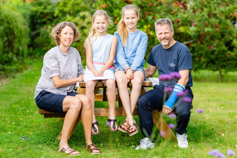 Familiefoto af smilende Virum familie på havebænk med grønne træer i baggrunden, naturligt lys.